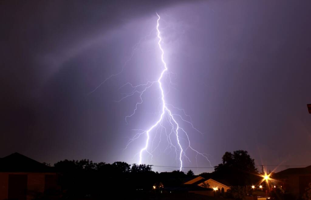 Color night time photo of lightning against a cloudy dark purple sky landing near a house.
