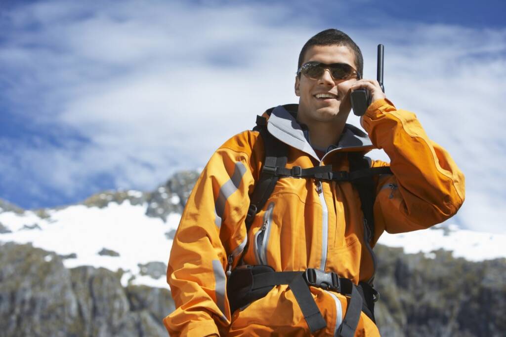 Color daytime photo of a young brunette man holding and talking on a satellite phone on his left ear. Partly cloudy blue sky with high altitude mountain background.