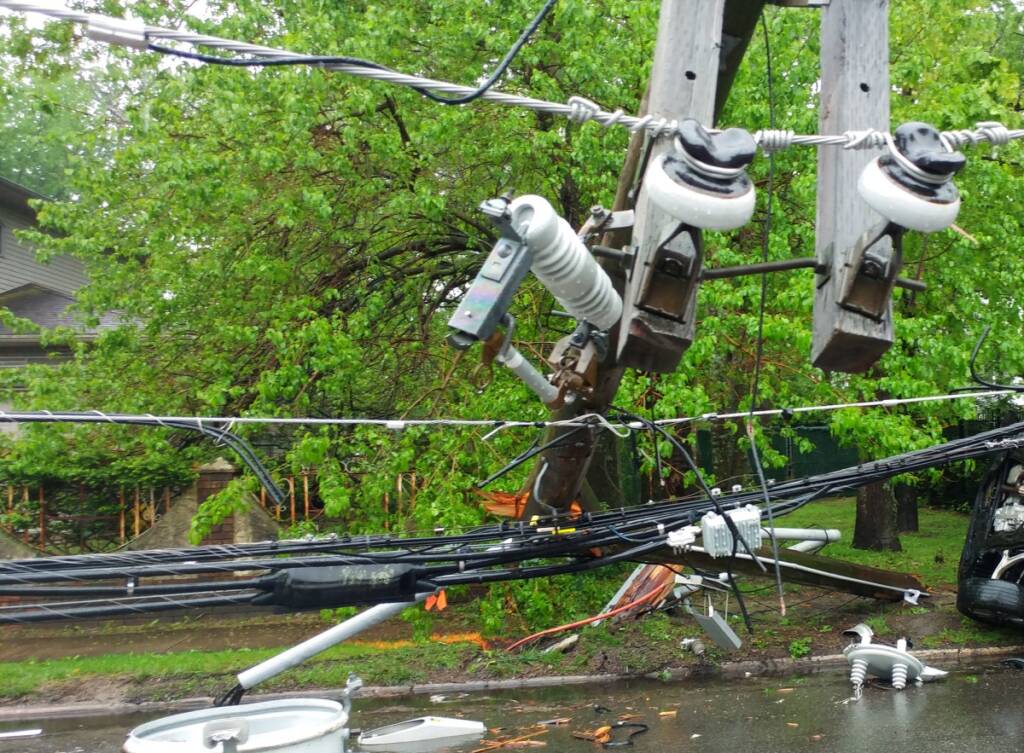 Color daytime photo of powerful storm damaged electric transformer on a pole laying in the street and a wet rain soaked trees damaged.