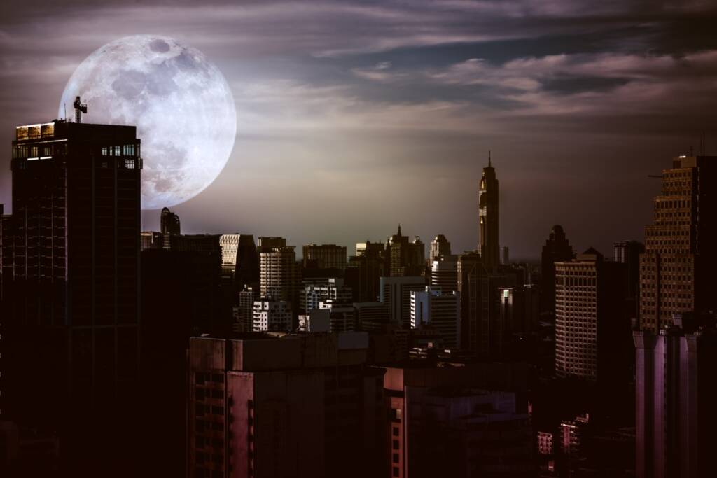Night time color photo of a large ful moon backdrop of a metropolitan city with silhouettes of dark skyscrapers in a total electrical power black out caused by a G5 Geomagnetic Carrinton Event Level Storm.