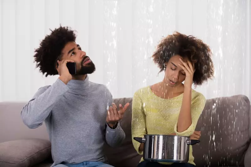Color indoor photo of a young black man using his cell phone calling for help in a flood from his ceiling while his wife is collecting the water leaking in a pan.