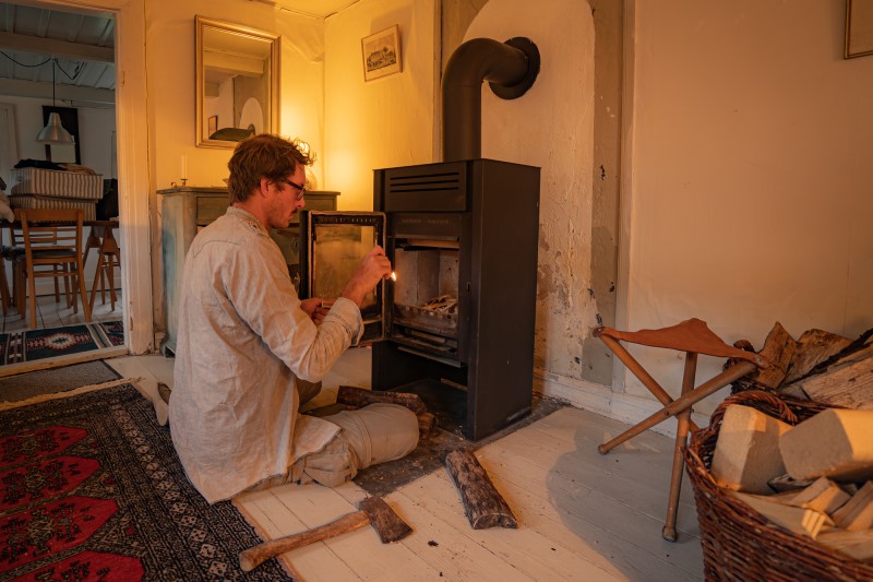 Color photo of a young white man lighting a wood stove to heat his home.