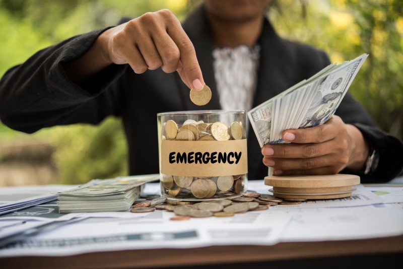 Color photo of a black woman's hand closeup holding 100 dollar bills in one hand and putting coins in a full emergency labeled clear glass jar.