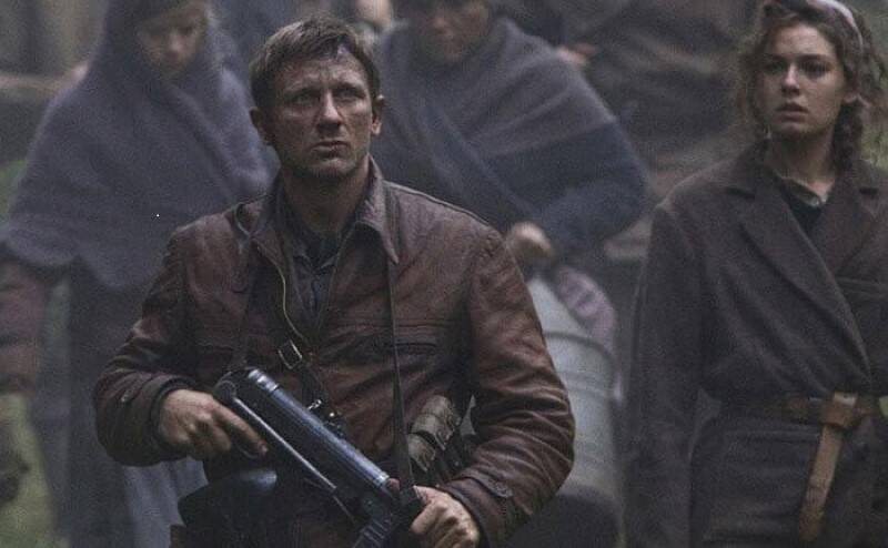 Color daytime photo of the Actor Daniel Craig holding a machine gun and playing one of the Polish Jewish Bielski brothers in the movie Defiance in front of a group of women.