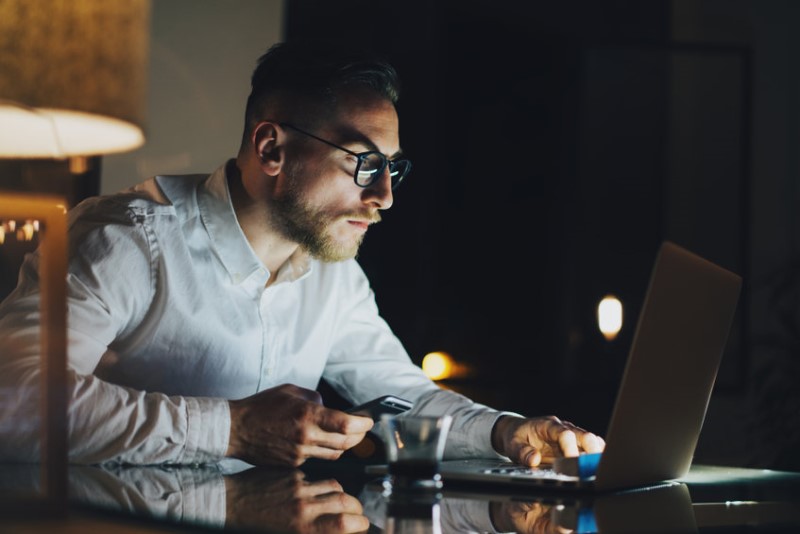 Color photo of a young man with glasses working with a laptop and smartphone at night observing remote technology.